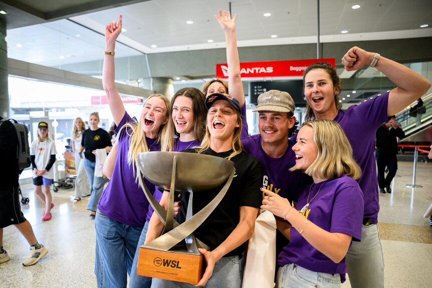 Surfing champion Molly Picklum holds trophy, posing with famil and friends, at airport