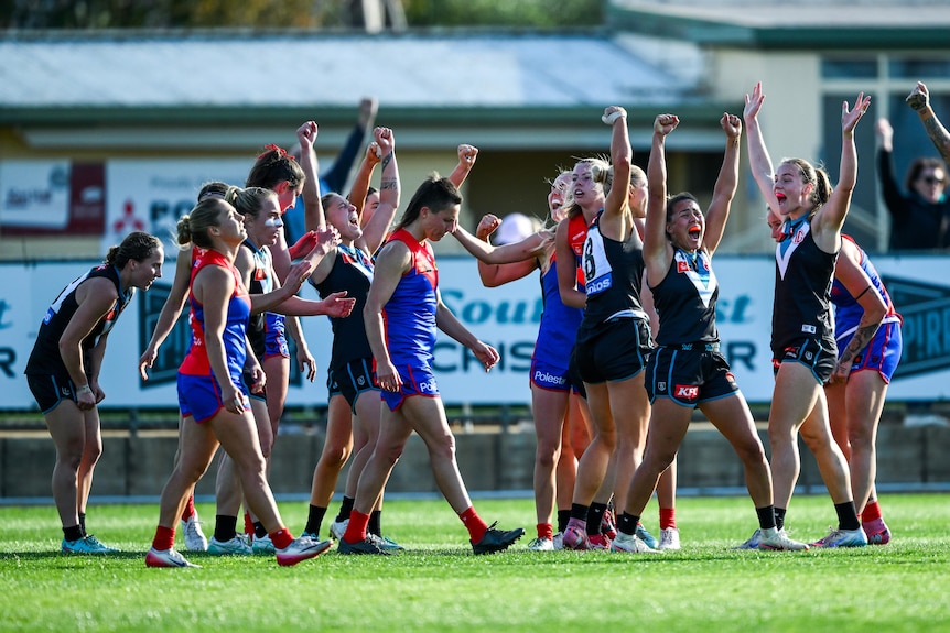 A group of Port Adelaide AFLW players cheer and raise their arms in celebration, surrounded by dejected Melbourne players.