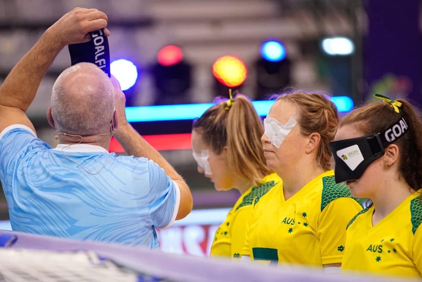 Three women in yellow and green stand in a line with eyeshades on next to a man in a blue shirt holding a black piece of cloth