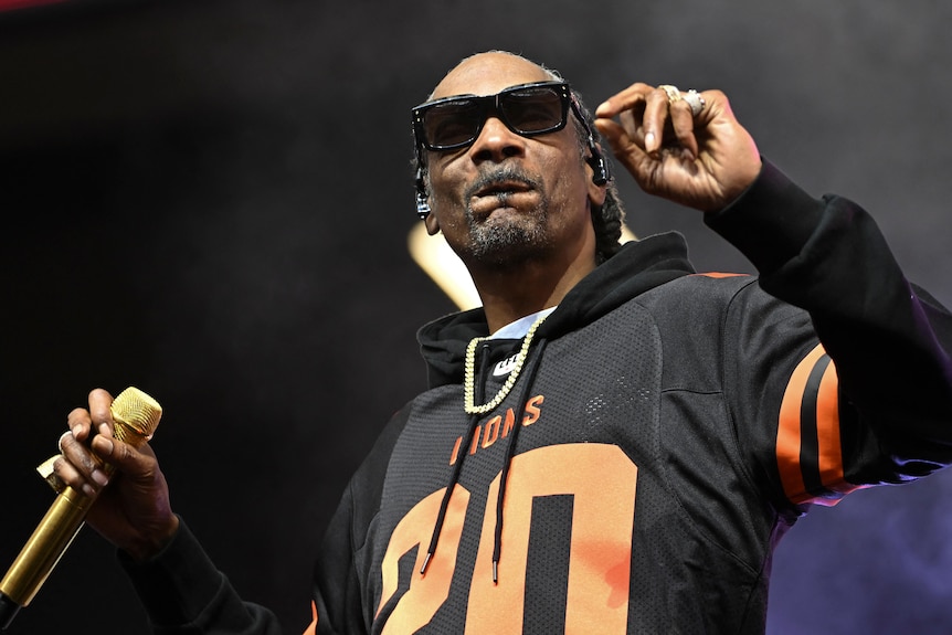 Snoop Dogg on stage in a black hoodie, wears sunglasses, holds a mike, other hand is at shoulder height, as if clicking fingers.