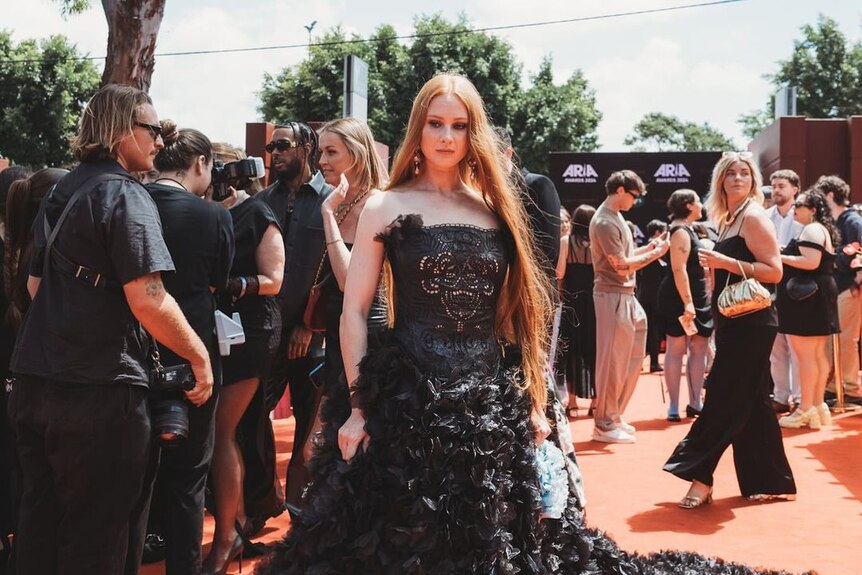 Vera Blue in a gothic gown stands in front of a large crowd of people in an open area surrounded by trees.