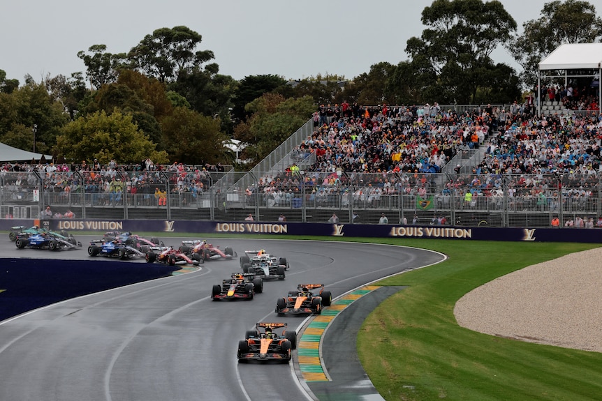 Ten Formula 1 cars snake around a bend on a track on a rainy day near a full grandstand