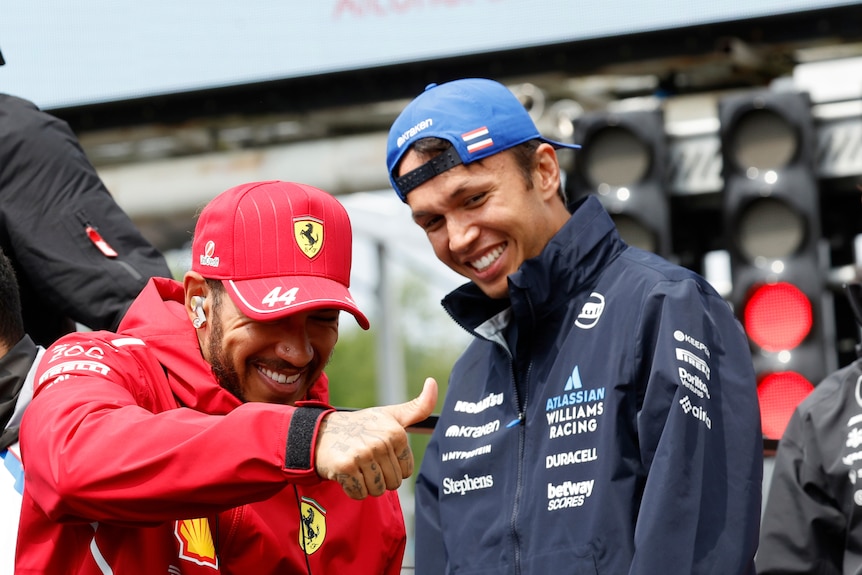 A man dressed in a red Ferrari cap and jacket smiles and gives the thumbs up with another man