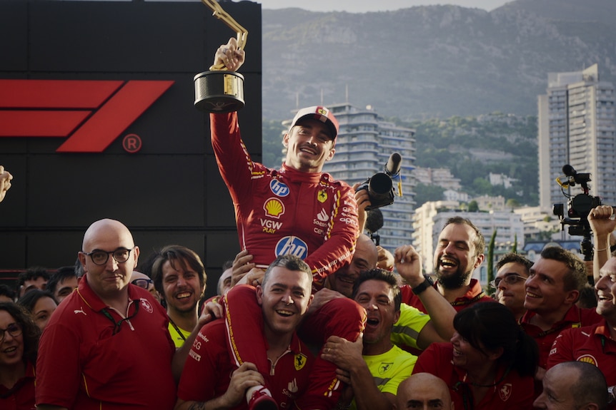 A Formula 1 driver dressed in Ferrari red holds up a trophy as he sits on a man’s shoulders