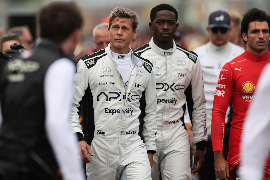 Actor Brad Pitt is wearing a white Formula One team racing suit, his co-star Damson Idris walks behind him.