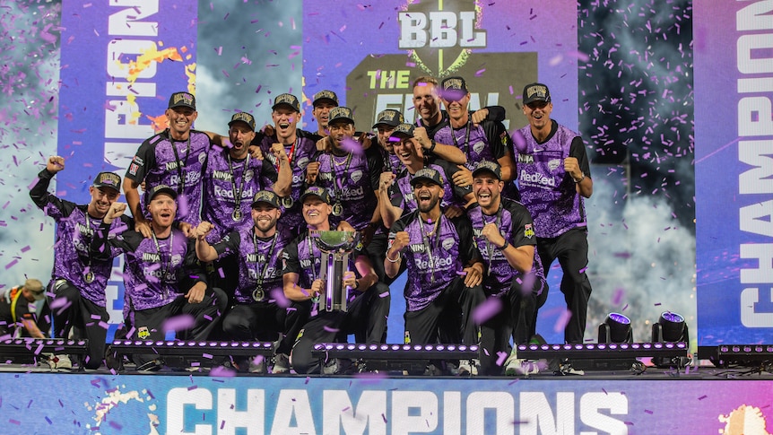 A group of Hobart Hurricanes cricketers stand on a podium and celebrates while holding a trophy as confetti falls around them.