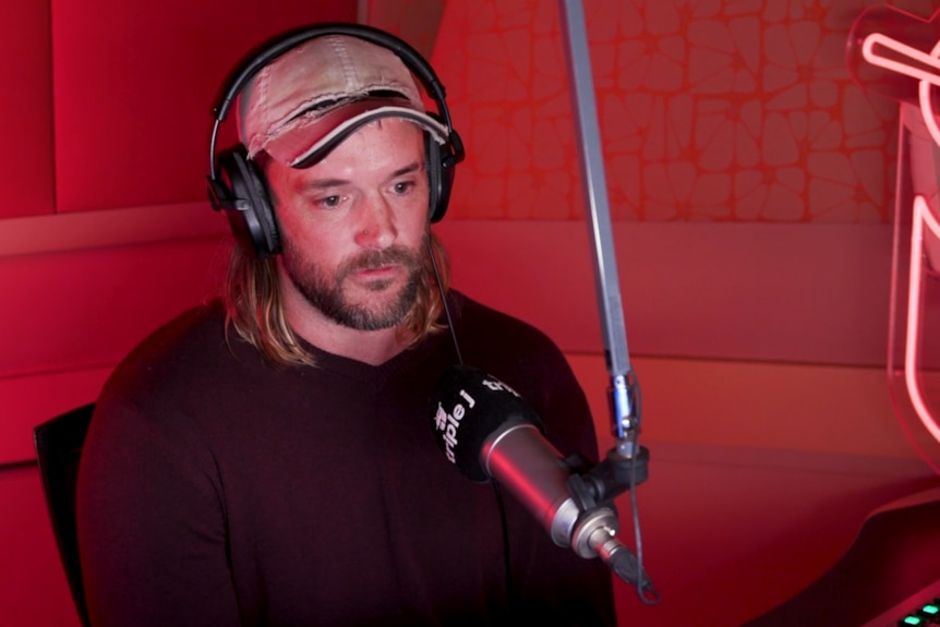 Man has a serious expression as he sits in front of a radio microphone. He has shoulder length brown hair and wears a cap. 