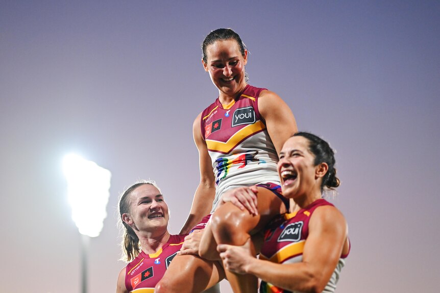 An AFLW player is lifted on the shoulders of two other players, all in red and white