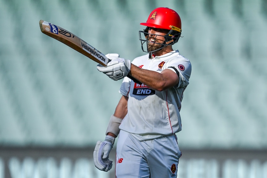 A cricket player in whites and red helmet holds up his bat in celebration of century