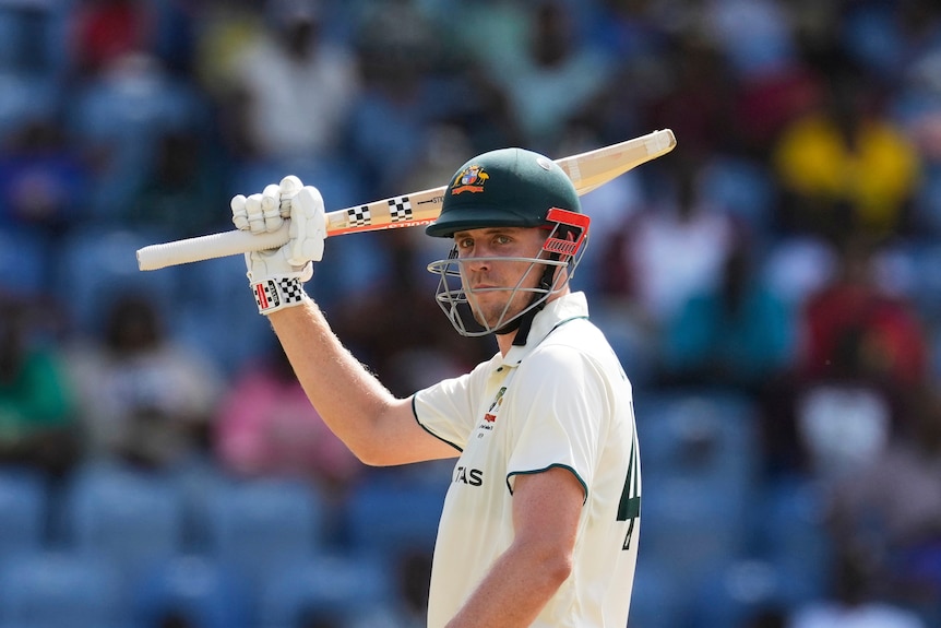A cricket player with green helmet raises his bat in front of a crowd.