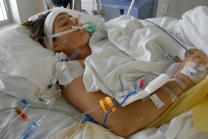 Katherine at 19 lying in a hospital bed with breathing apparatus in her mouth, and numerous tubes running to her arms and body.
