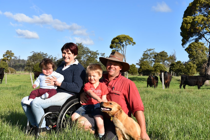 Katherine, in her wheelchair, with a man and two small children in a paddock full of green grass. Cattle graze nearby.