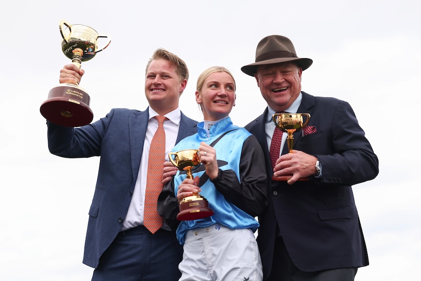 A jockey stands between two men at Flemington and looks up at the smiling man next to her as they all hold race trophies.