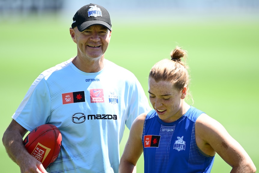 A smling man in a cap holds a football while standing next to a female AFL player on an oval.