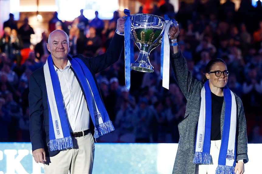 A man and a woman, both wearing football scarves, hoist a large trophy in front of a crowd.