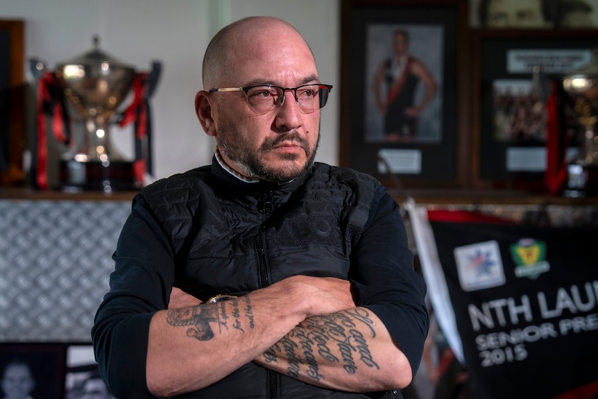 A man with glasses and sleeve tattoos sits with arms folded and trophies and awards in the background.