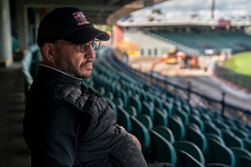 A man in a black cap and glasses sits in a stadium with turquoise seats and construction in the background.