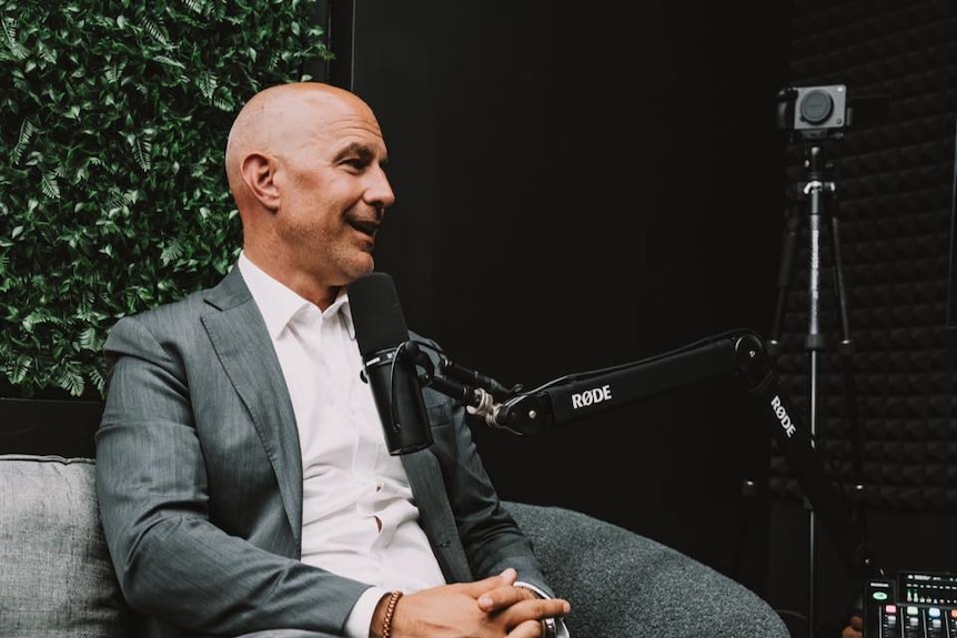 A man in casual business attire sits on a couch and talks into a podcast microphone