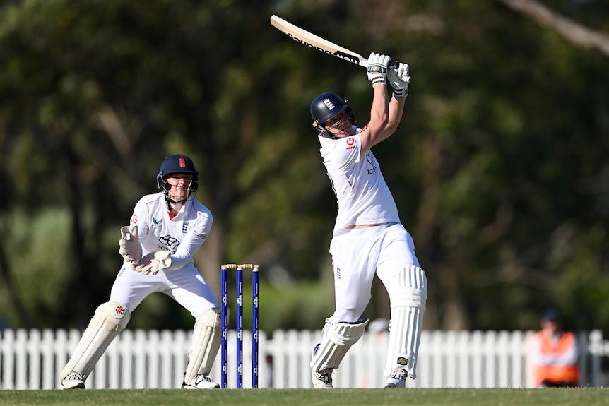 An England batsman extends his arms as he smashes the ball over the boundary in a tour game, as a wicketkeeper looks on.