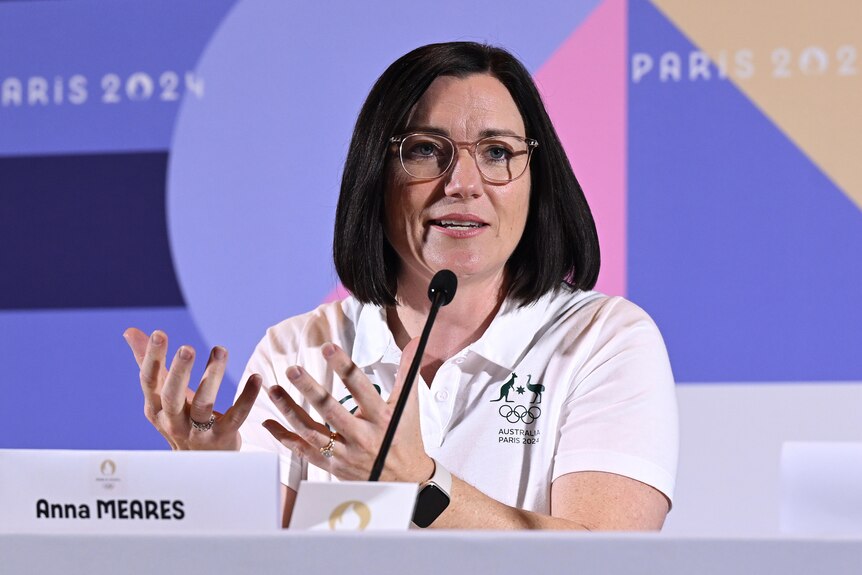 A woman wearing an Australian Olympic team shirt gestures as she speaks into a microphone at a press conference.
