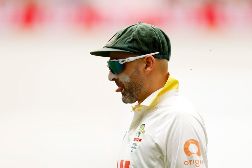 A cricketer in white and with a green hat looks down and pokes his tongue out