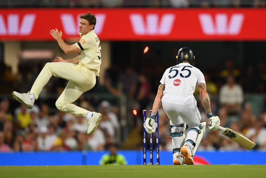 Brendan Doggett jumps as the stumps of Ben Stokes are hit by a ball. Stokes has his back turned.