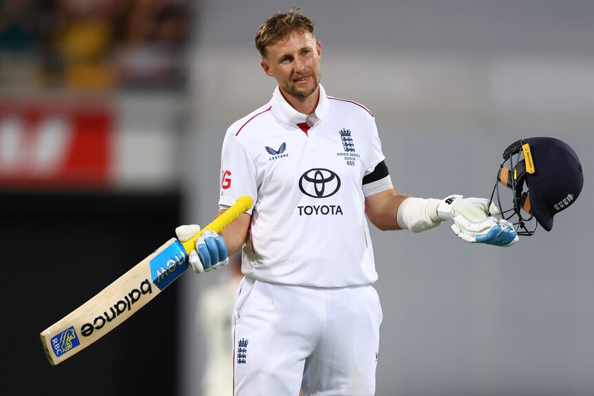 A cricket player in white holds up his helmet and bat while smiling