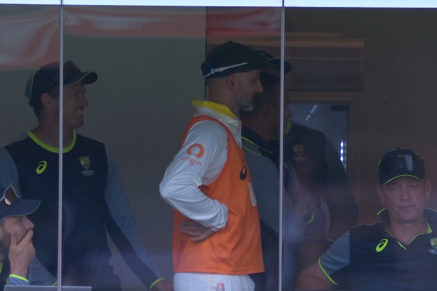 A man in a high-vis orange singlet over cricket whites stands in a changeroom with hands on hips