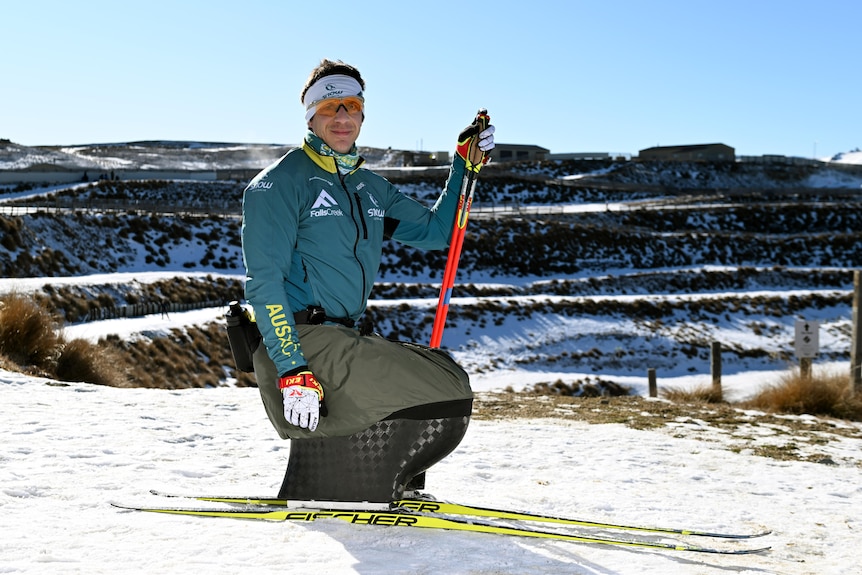Dave is smiling, sitting on his sit ski in a snowy field, holding his ski poles in his left hand