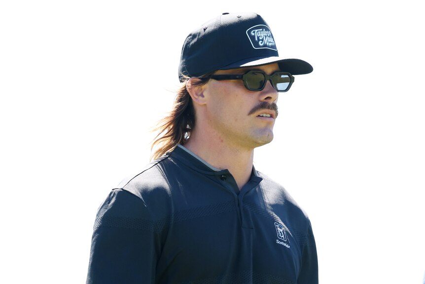 A young man with long hair and a moustache wears a dark cap, sunglasses and polo shirt.