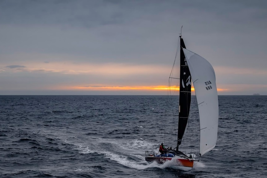 A yacht with black and white sails sailing in front of a sunrise.