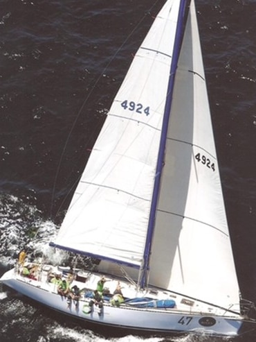 A boat is pictured from above with all-white sails and some crew sitting on the side as they sail during the Sydney to Hobart.