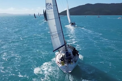 An aerial drone shot from behind of a race boat with sail up taking part in Airlie Beach Race Week.