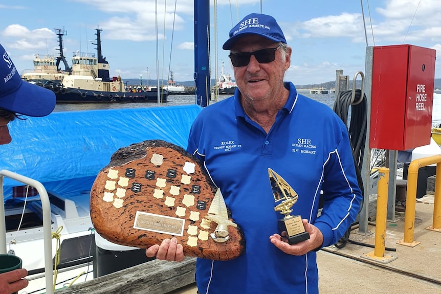 A boat captain stands on the dock at Eden in NSW holding two trophies, the bigger one a wooden one.