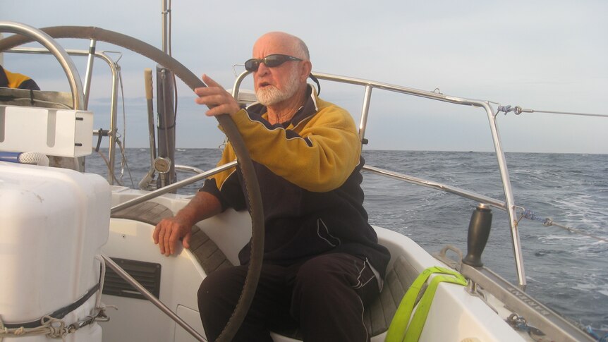 A sailor sits at the stern of his boat with his hands on the wheel while at sea.