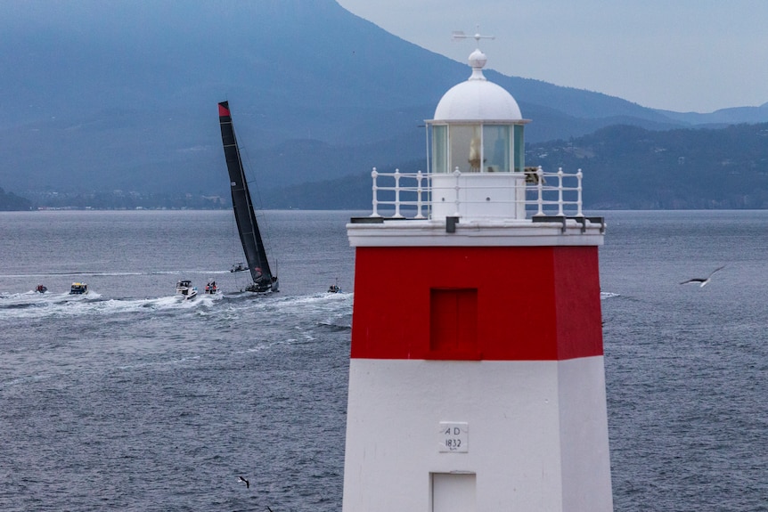 A view of the top of a red and white lighthouse looking down a river, with a super maxi and other boats in the background. 