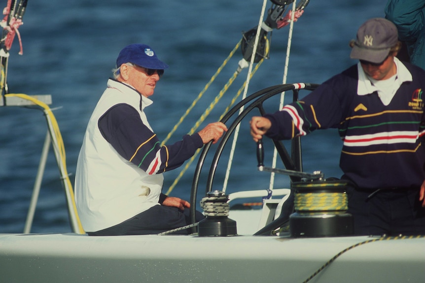 Australian sailing legend Syd Fischer holds the helm (wheel) of his America's Cup boat with a crew member beside him.