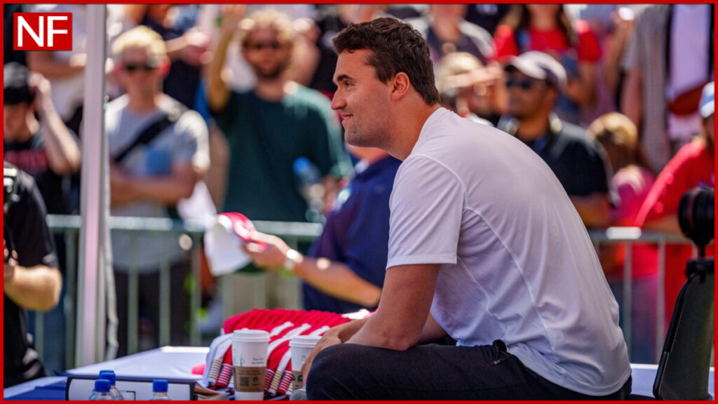 Charlie Kirk sits at a table in a white T‑shirt, facing a large outdoor crowd at Utah Valley University, with supporters and cameras gathered closely around him during a daytime event. 