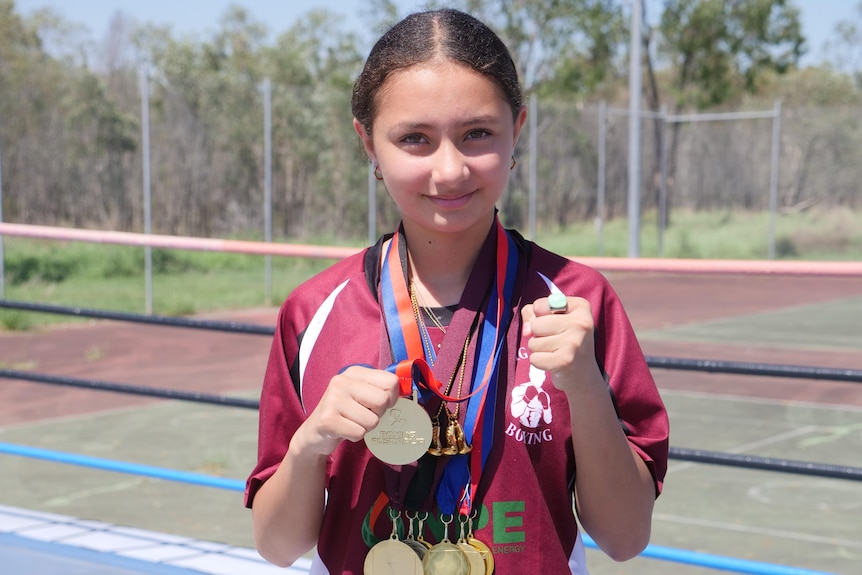 A tween girl standing in a boxing ring, holding medals and looking at the camera.