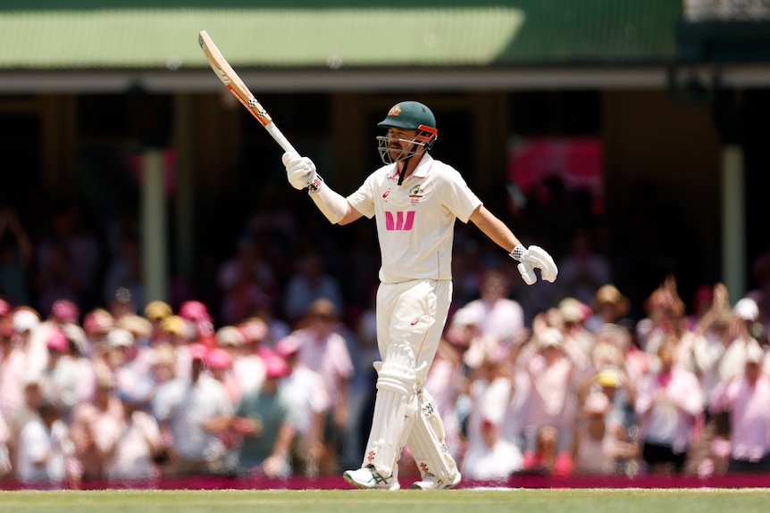 A cricket batter holds his bat up in celebration with crowd behind him