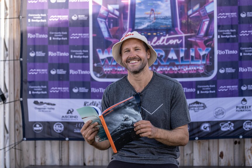 Image of Jaeger Stone holding a magazing and smiling. He has a short beard and wears a bucket hat and t shirt.