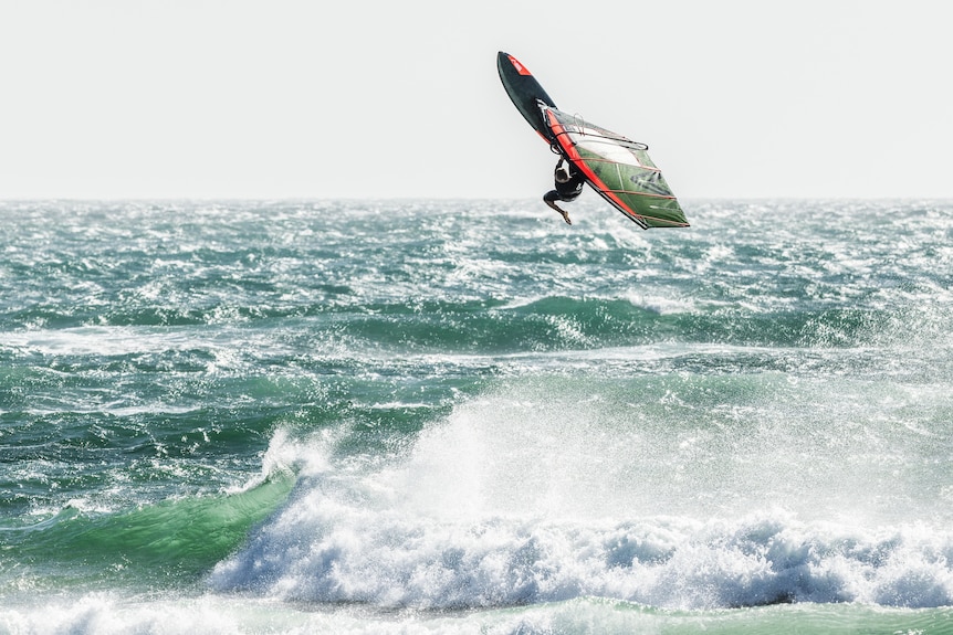 A windsurfer gets air above a wave and choppy ocean shoreline.