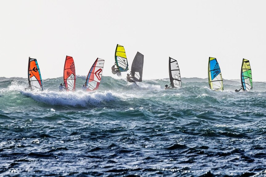 A line of windsurfers in the ocean. The waves are choppy and the sun is setting.