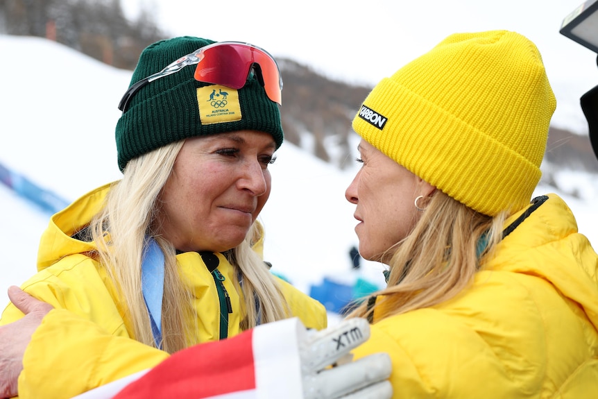 Two blonde women in beanies and parkas share a moment on a snowfield.