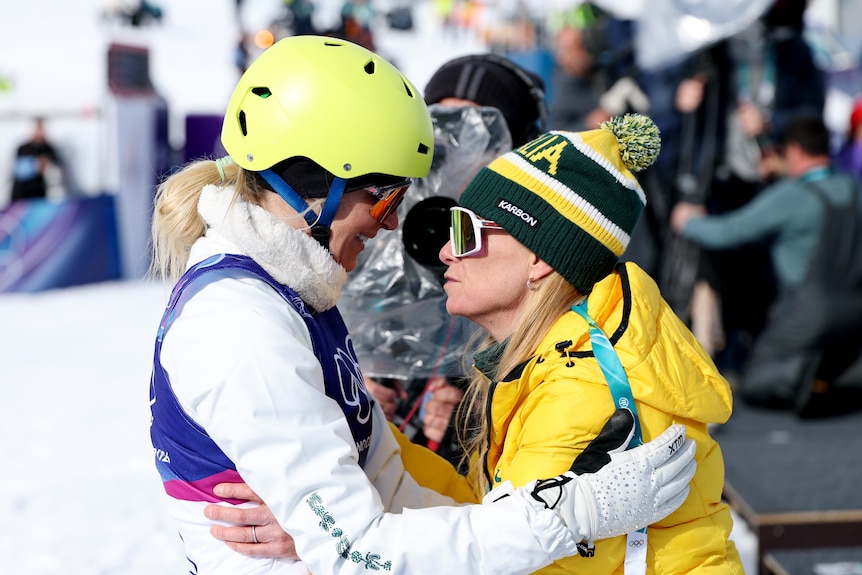A woman in ski gear and a helmet hugs a woman in a parka and beanie as they speak on a snowfield.