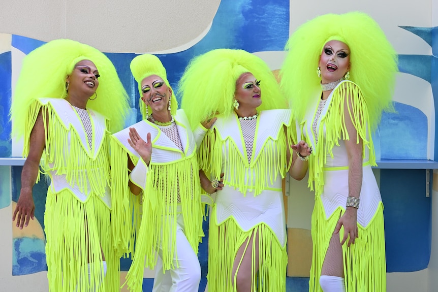 Four drag queens wear white outfits with neon yellow tassles, and bright neon oversized wigs