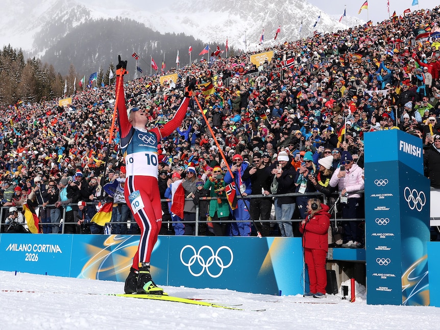 A Norwegian biathlete stands on his skis next to the finish line with his poles raised in triumph in front of a big crowd.
