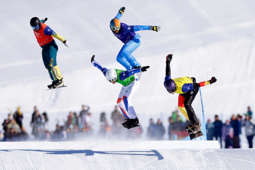 Adam Lambert snowboards in a race with three others, all in the air over a jump.