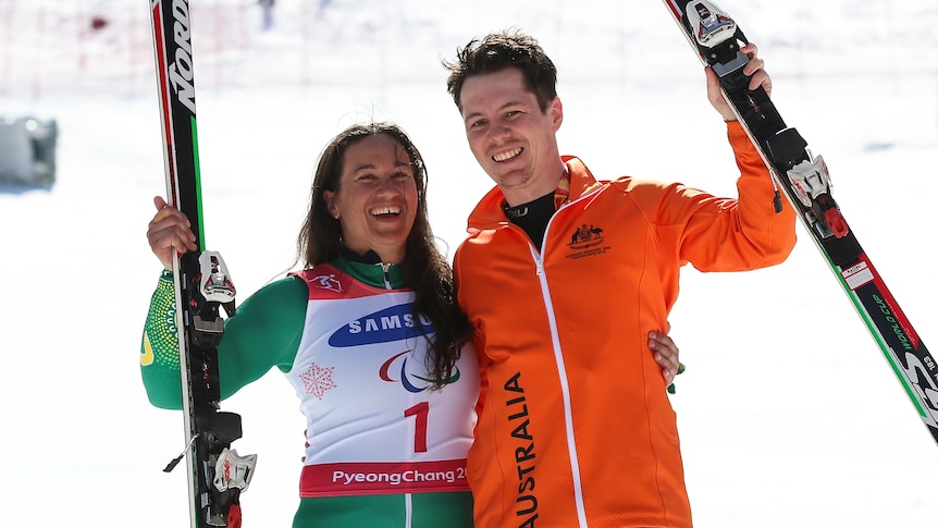 An Australian Paralympic skier and her guide stand smiling with their skis on the dais before receiving medals.