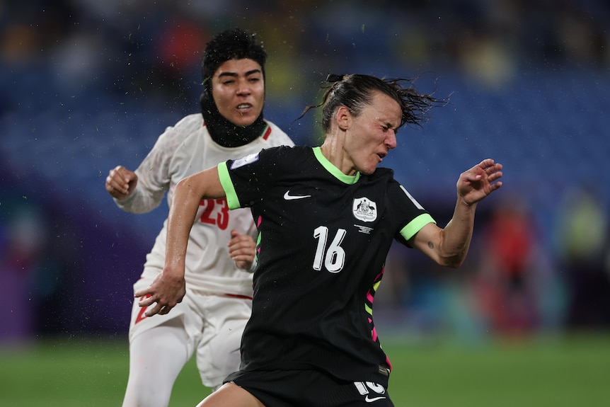 Matildas striker Hayley Raso grimaces after she is hit in the head by a hard-struck ball, with an Iran defender behind her.